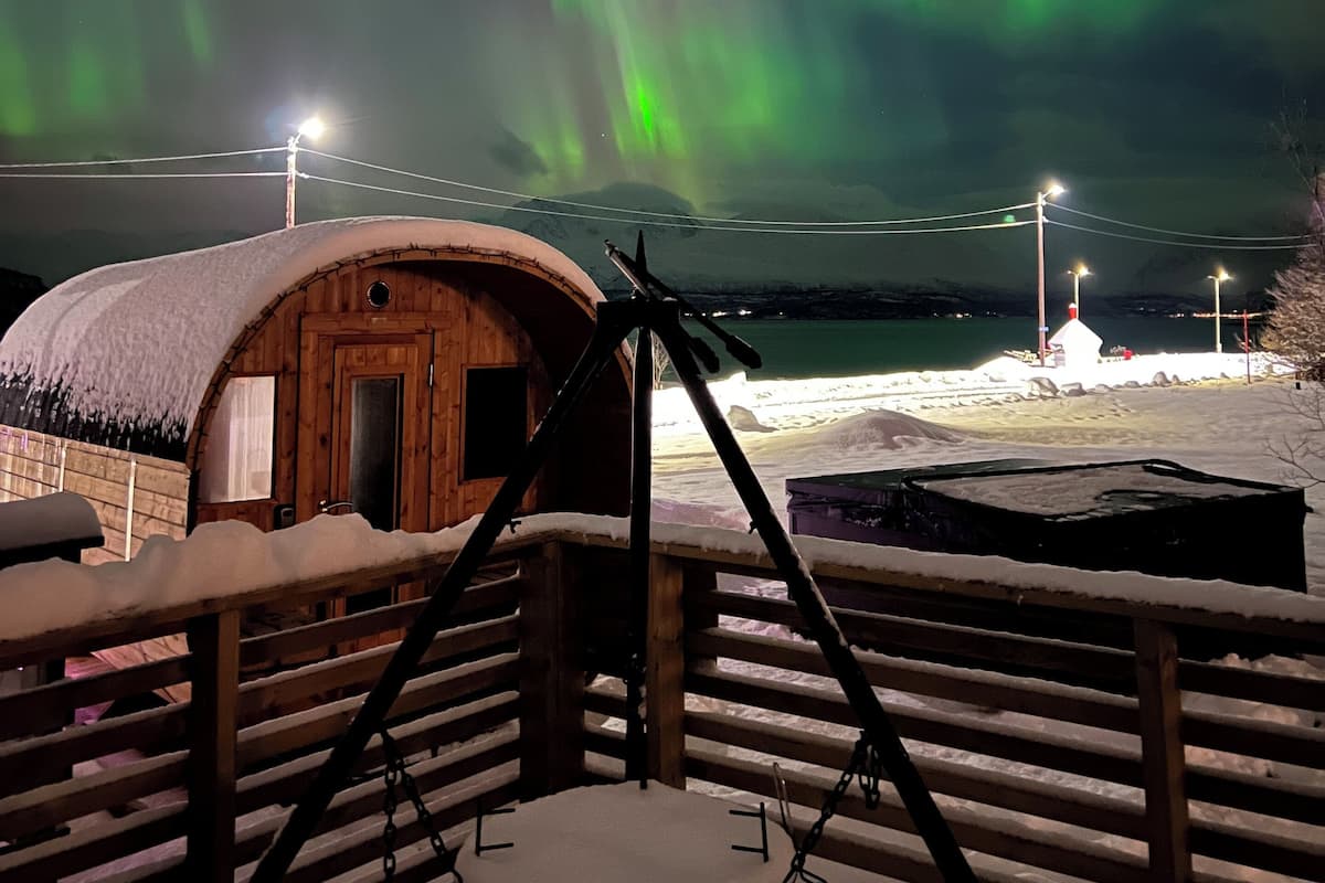 Una cabaña de madera con techo nevado, un telescopio y vistas de las auroras boreales.