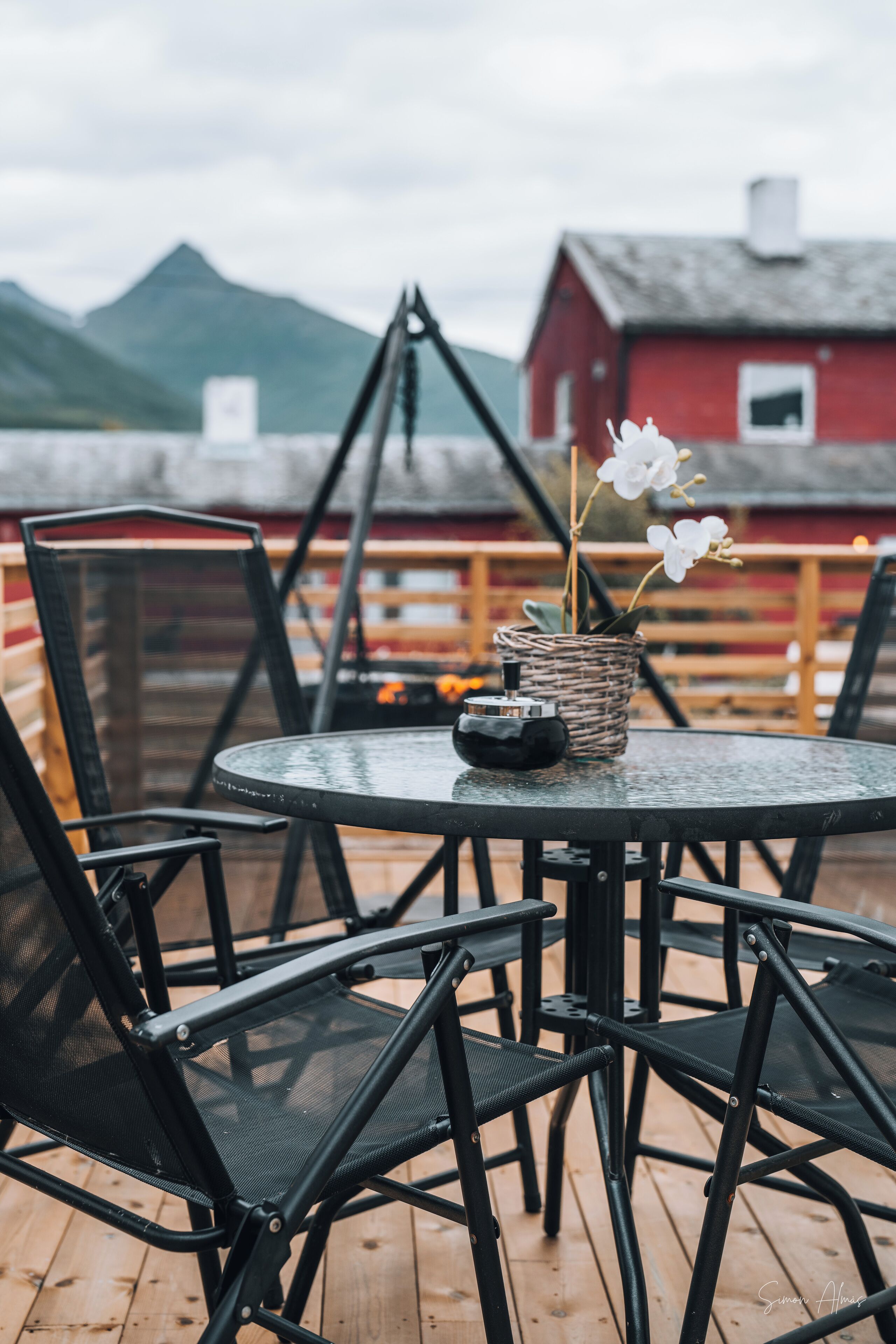 Una mesa redonda con sillas en una terraza de madera, una cesta de mimbre con flores y un edificio rojo al fondo.