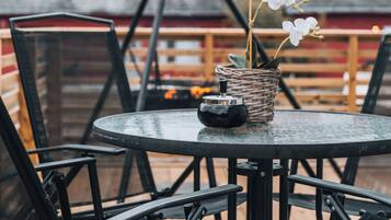 Una mesa redonda con sillas en una terraza de madera, una cesta de mimbre con flores y un edificio rojo al fondo.