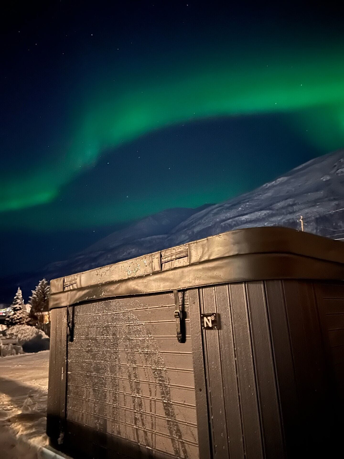 A wooden cabin with a metal door, set against a backdrop of a snowy landscape and the Northern Lights.