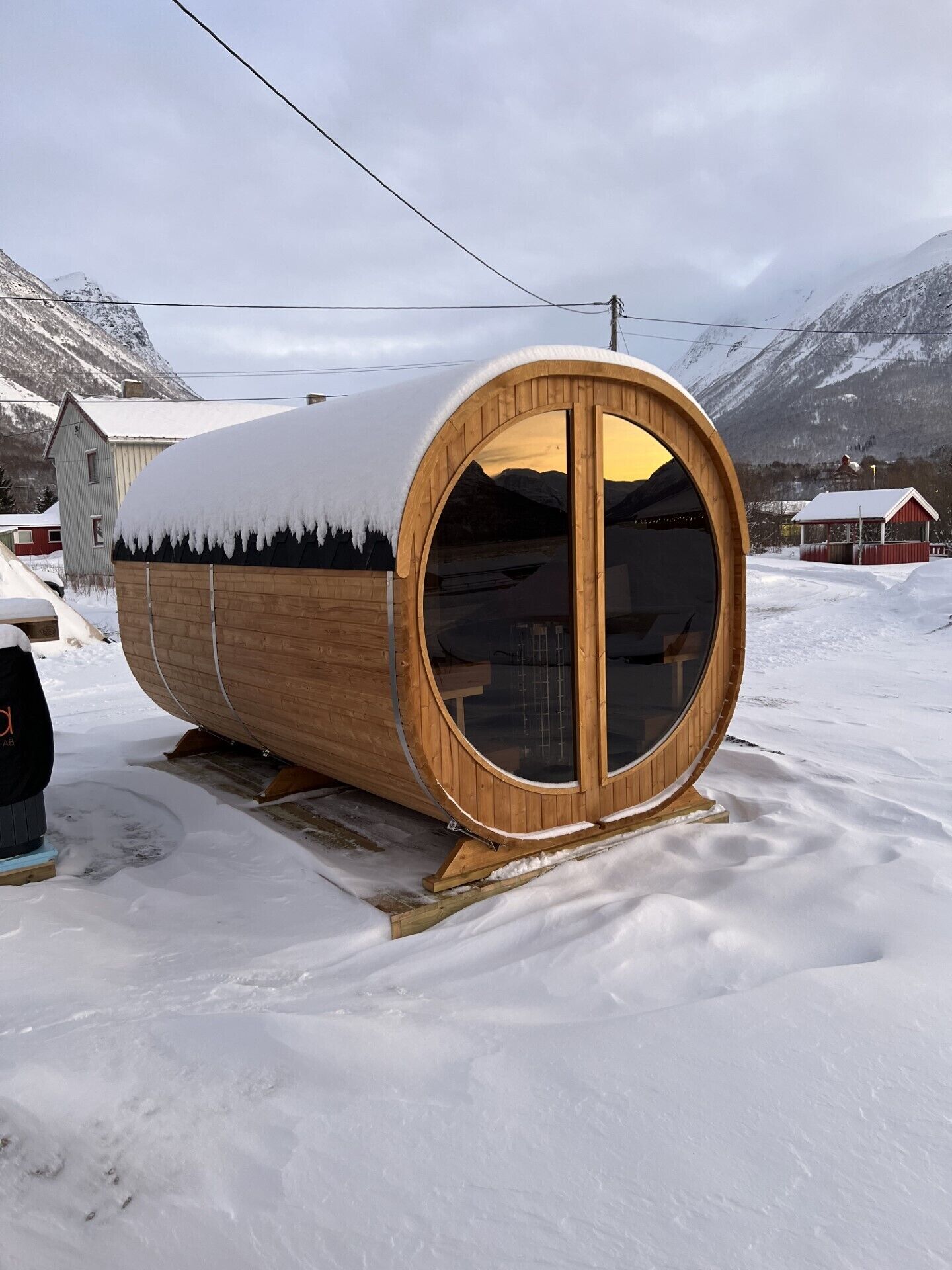 A wooden cabin with a large circular window, situated in a snowy landscape with mountains in the background.