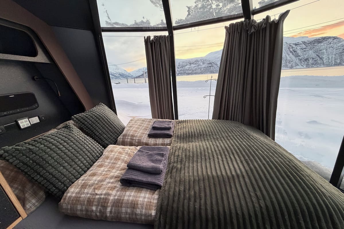 Interior acogedor de una cabaña con cama, almohadas y ventana que ofrece vistas a un paisaje nevado.