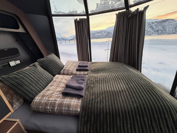 Een knus houten huisje van binnen, met een bed, kussens en een raam met uitzicht op een besneeuwd landschap.