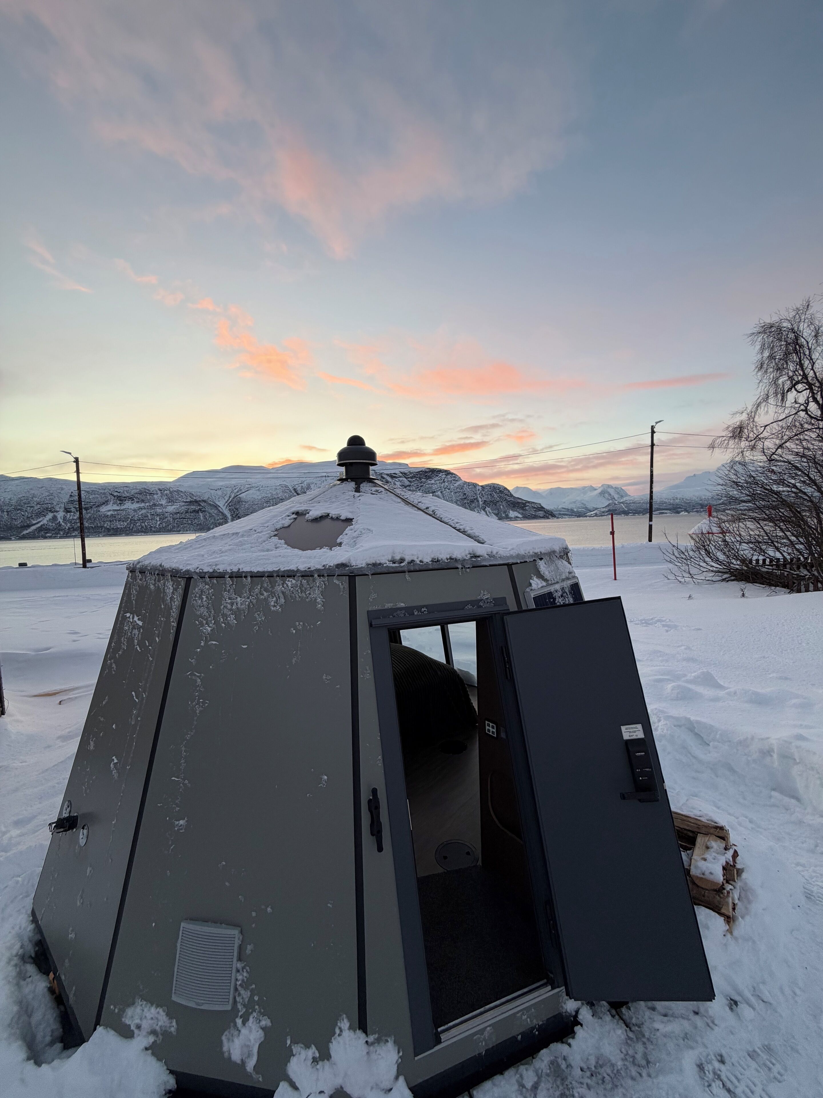 A snow-covered tent with a door open, set against a backdrop of mountains and a sunset sky.