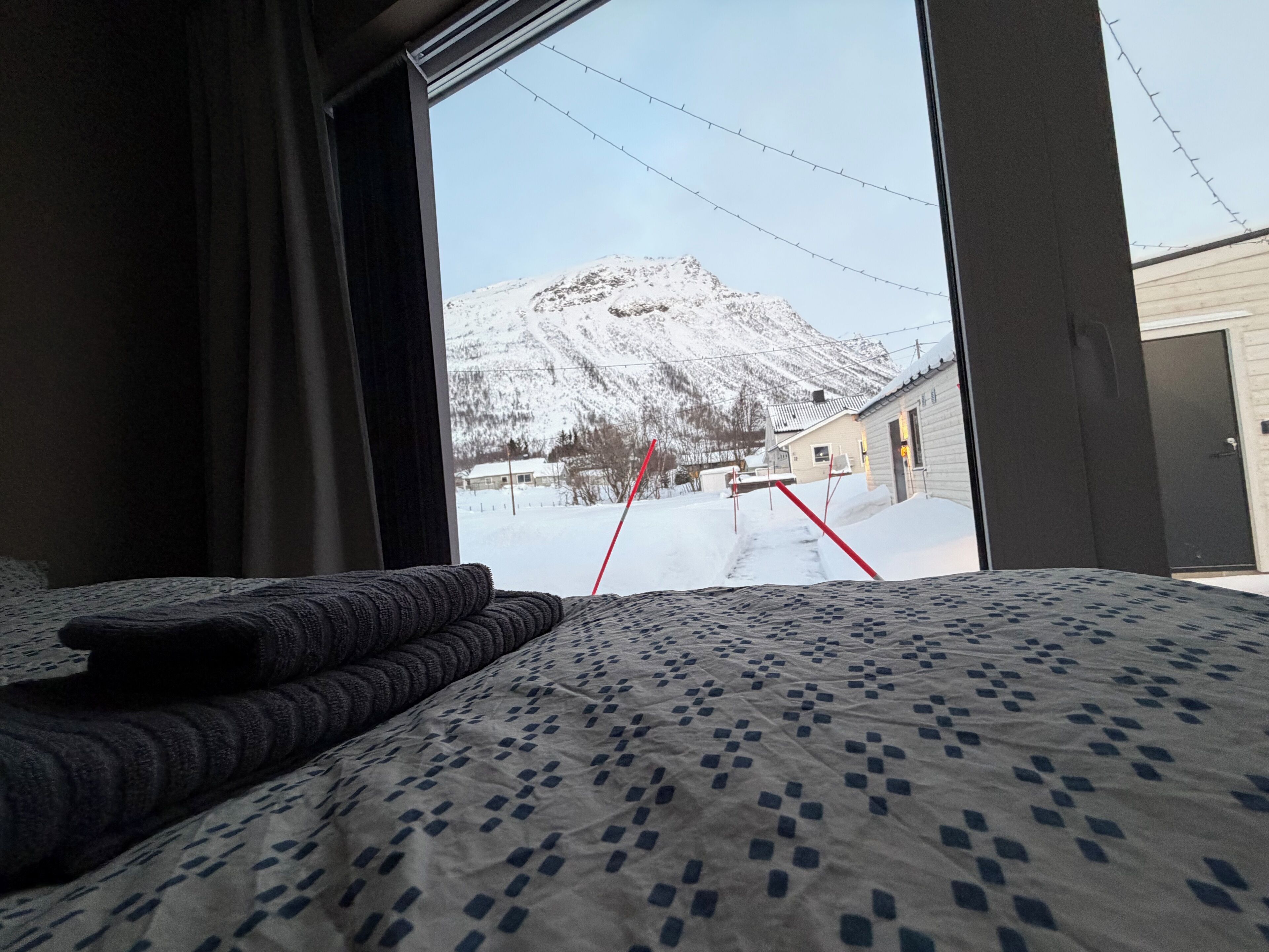 Ein Schlafzimmer mit einem großen Fenster, einer gemusterten Bettdecke und Blick auf einen verschneiten Berg sowie ein Gebäude in der Ferne.