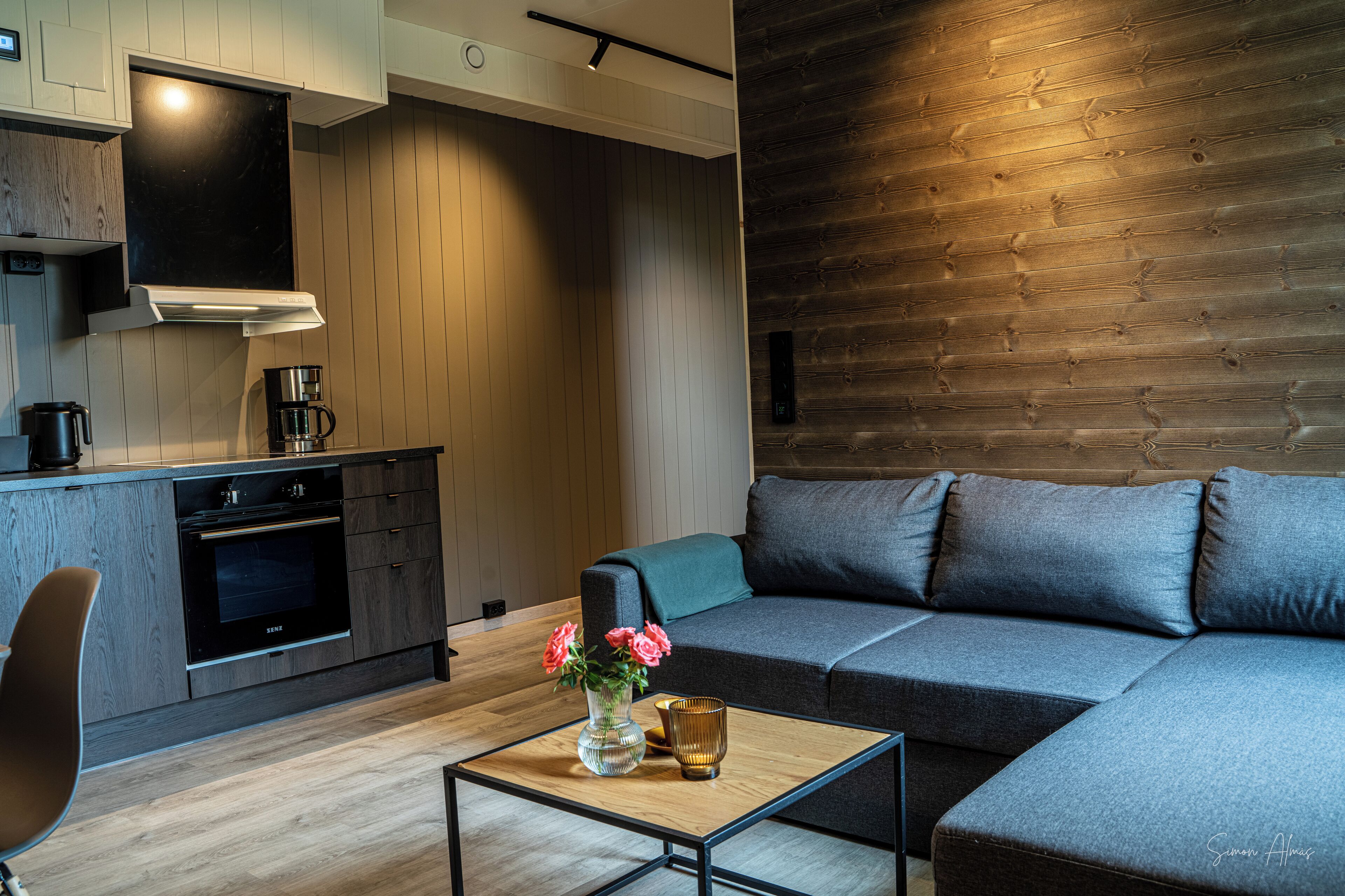 A modern kitchen with a built-in oven and a coffee machine, a grey sofa with cushions, and a small table with a vase of flowers.