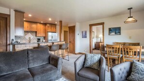 Ein modernes Wohnzimmer mit grauem Sofa und Sesseln, eine Essecke mit Holztischen und -stühlen sowie eine Küche mit Holzschränken und einem Edelstahlkühlschrank.