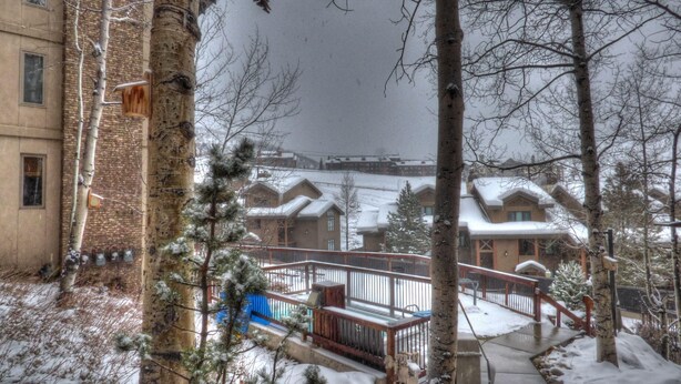 Ein verschneiter Außenbereich mit einer Holterrasse, einem Metallgeländer und einem Gebäude mit schneebedeckten Dächern im Hintergrund.