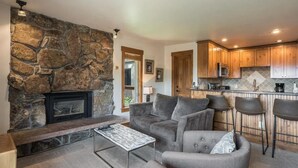 Ein Wohnzimmer mit Kamin aus Stein, grauen Sofas und einer Küchenzeile mit Holzschränken und einer Mikrowelle.