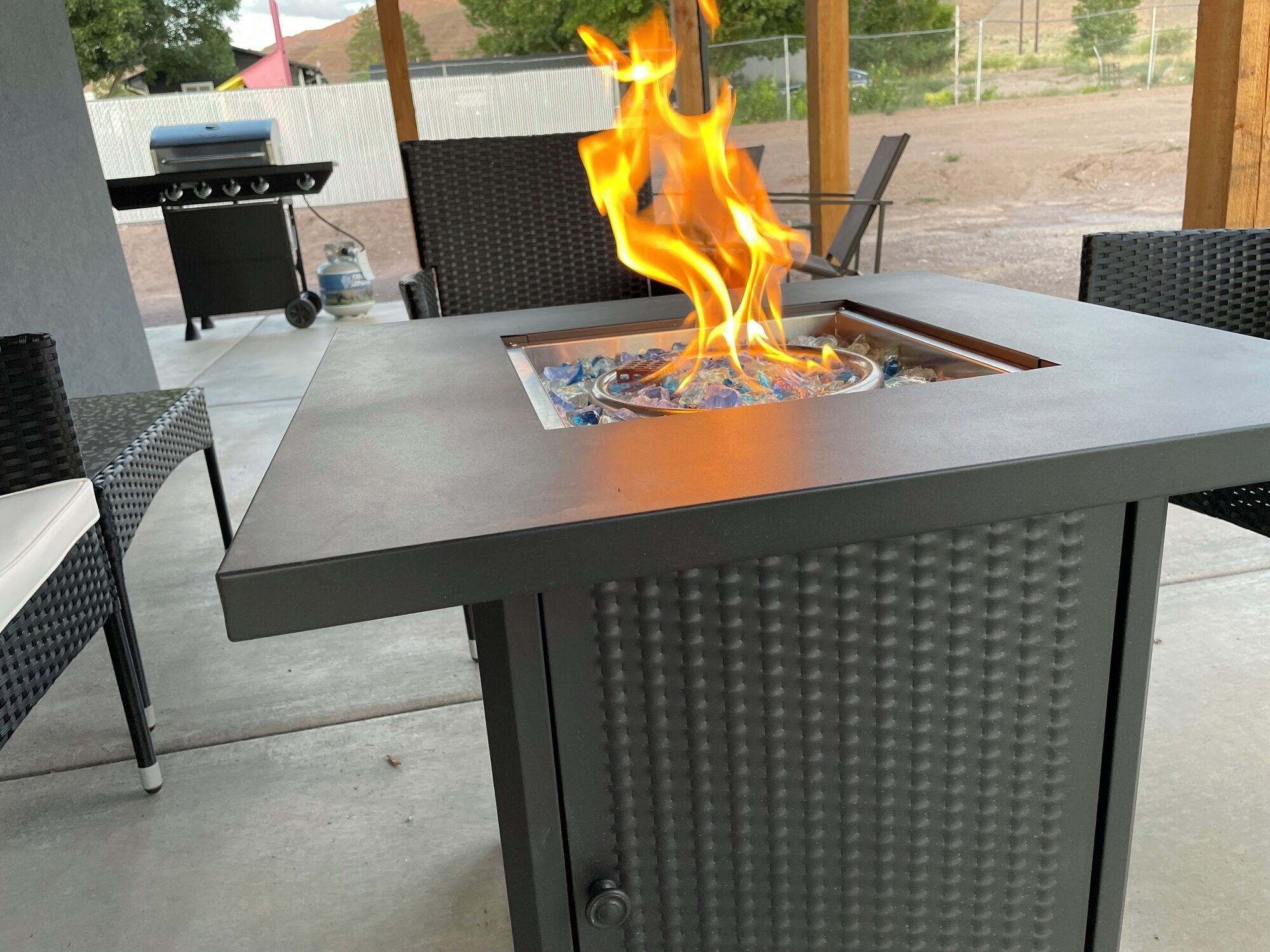 Une terrasse extérieure en métal, dotée d’un foyer intégré, de chaises en osier et d’un barbecue à l’arrière-plan.