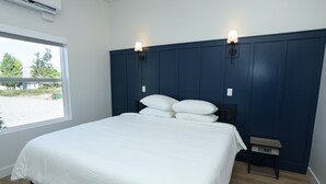A bedroom with a large bed, two wall-mounted lights, a small bedside table, and a window with a view of greenery and buildings. - Mini Cottage Rentals, Monroe UT. Come Ride the trails (Monroe)