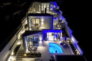 A modern multi-level house with a swimming pool, outdoor seating, and a well-lit interior. - White Cliff Luxury Suites (Markopoulo Mesogaias)