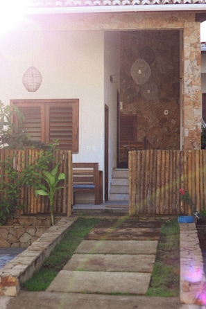 A house with a stone facade, a wooden door, and a small garden area with a bench and plants. - Pousada Casa de Hóspedes (Amontada)