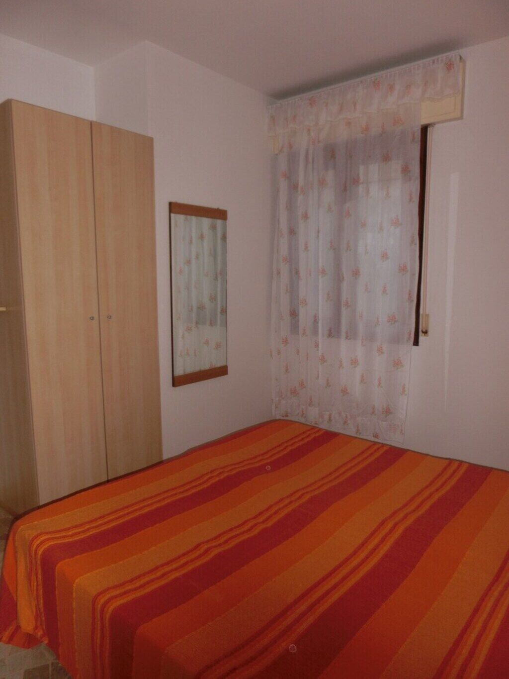 Een slaapkamer met een bed, een kledingkast, een spiegel en een raam met bloemenkurten.
