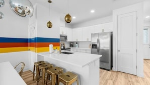 Cocina moderna con armarios blancos, refrigerador de acero inoxidable, una isla central con taburetes y una franja de colores en la pared.