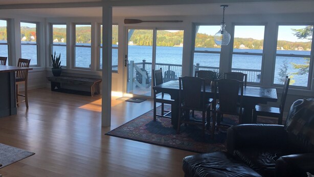 Ett rymligt vardagsrum med ett stort fönster med utsikt över en sjö, en matplats med fin utsikt och en mysig sittgrupp med en lädersoffa.