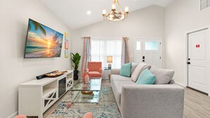 Een moderne woonkamer met een bank, fauteuils, een salontafel en een televisie. Er is een kroonluchter, een plant en een deur met een bordje.