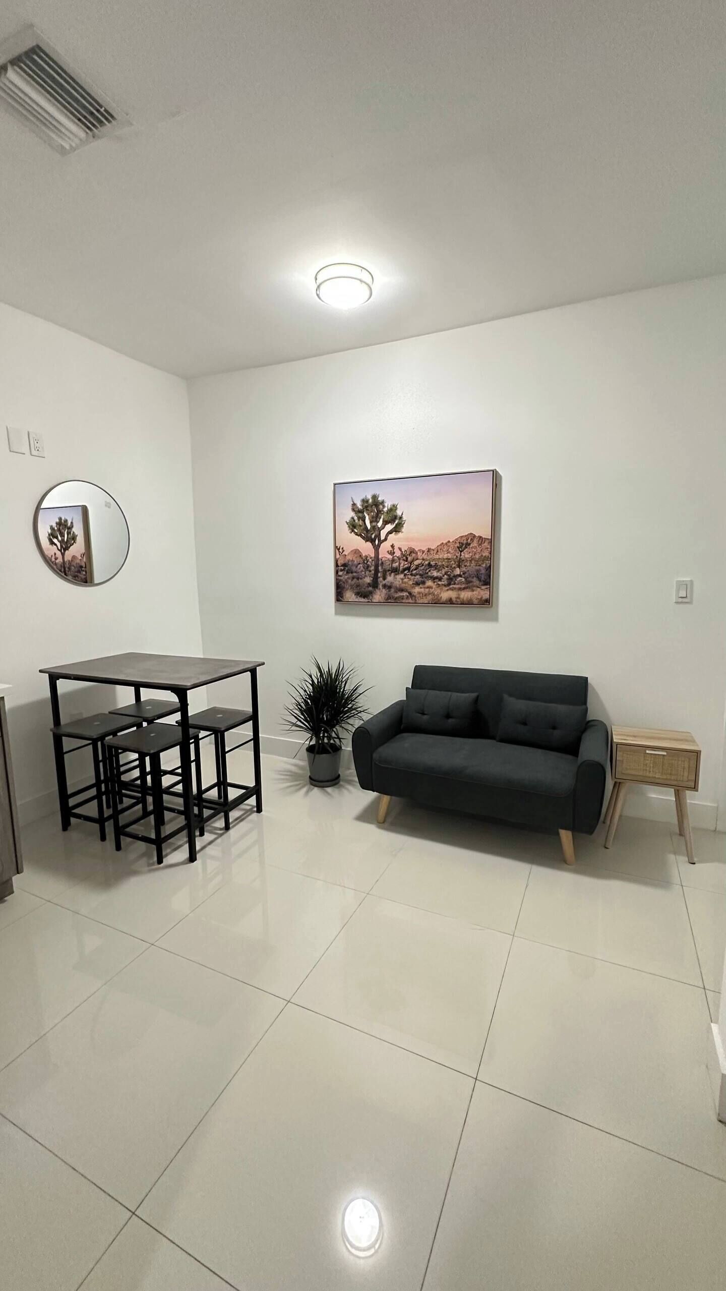 Una sala de estar moderna con un sofá negro, una mesa de comedor con sillas, una planta en maceta y un cuadro colgado en la pared.