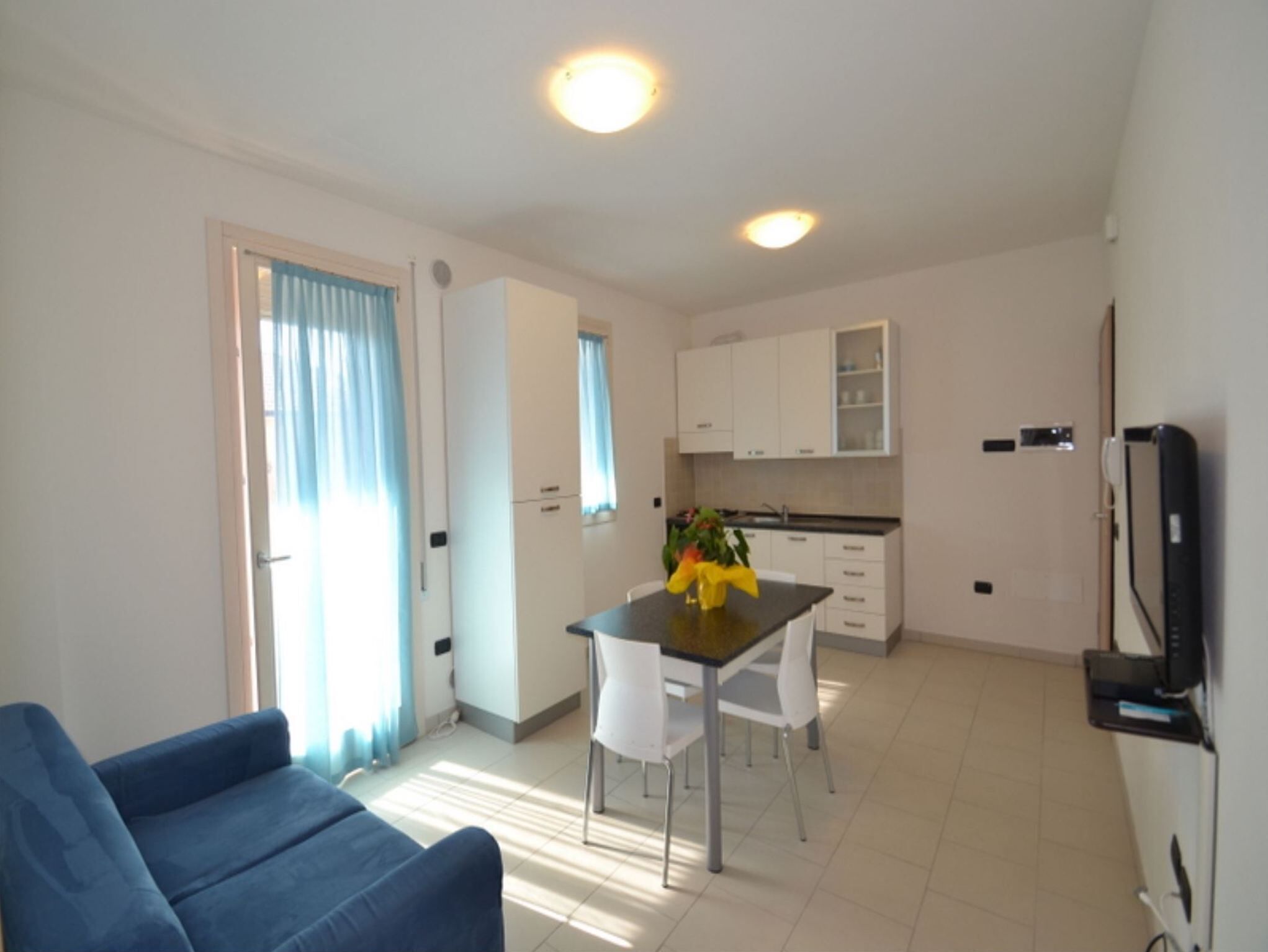 A modern living room with a blue sofa, a dining table with chairs, a kitchen area with white cabinets, and a TV mounted on the wall.