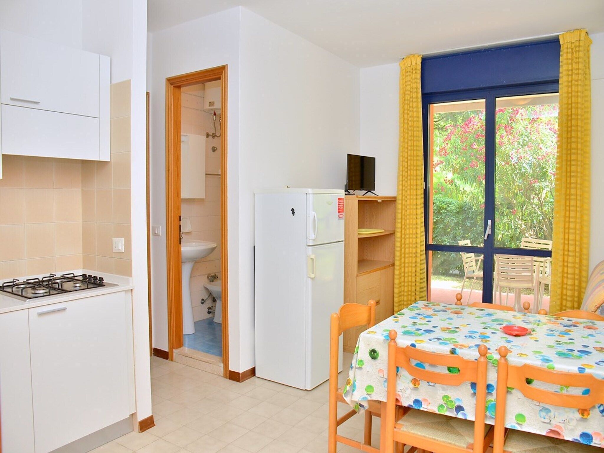 Cozinha compacta com fogão a gás, geladeira e pia. Há uma pequena área de refeições com mesa e cadeiras, e uma janela com cortinas amarelas.