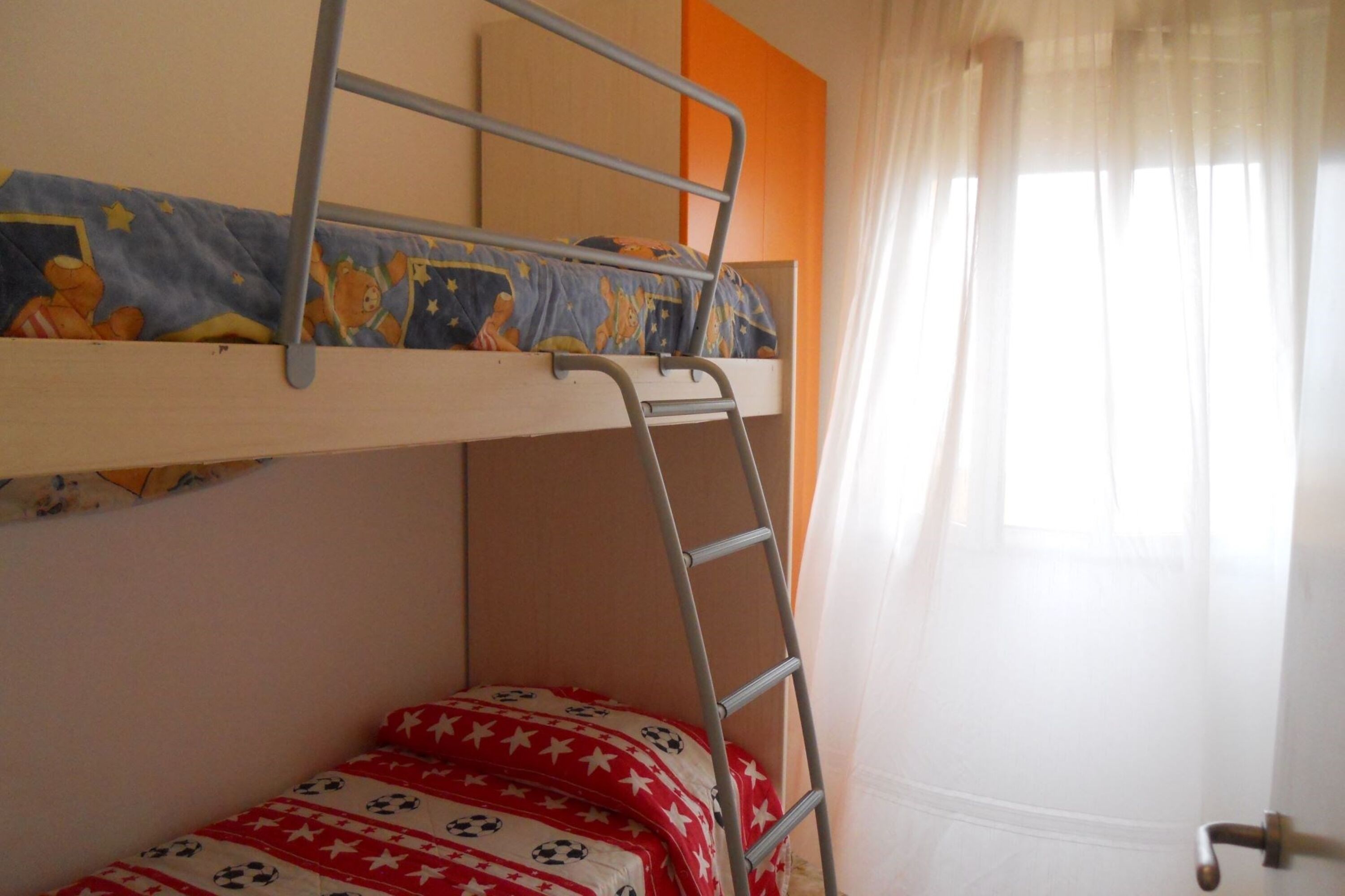 Un llit superposat amb una escala, una finestra amb cortines lleugeres i una porta amb manilla.