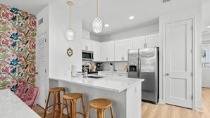A modern kitchen with white cabinets, a stainless steel refrigerator, a central island with stools, and pendant lighting. - Welcome to Paradise! ~ STEPS to BEACH! ~ Sleeps 10 (Panama City Beach)