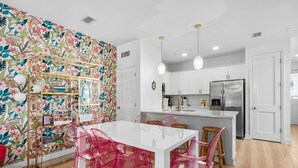 A modern kitchen with white cabinets, a stainless steel refrigerator, and a dining area with a white table and pink chairs. - Welcome to Paradise! ~ STEPS to BEACH! ~ Sleeps 10 (Panama City Beach)