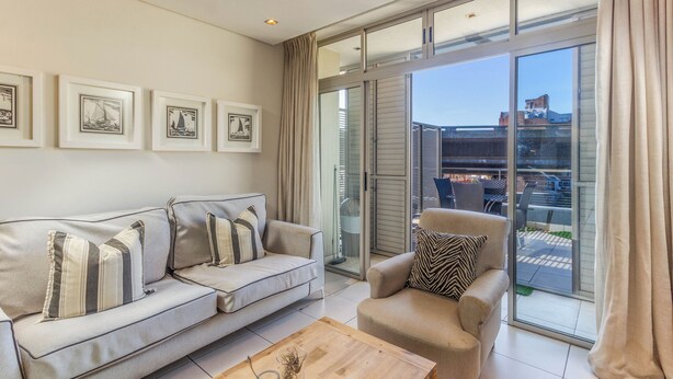 Una sala moderna con sofá, sillón y mesa de centro, puertas de cristal correderas hacia un balcón y obras de arte enmarcadas en la pared.
