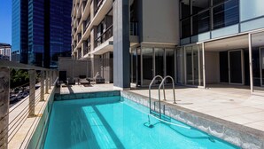 Una piscina en la azotea con una escalera de metal, rodeada de edificios modernos y un cielo despejado.