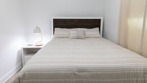 A neatly made bed with a striped bedspread and pillows, a bedside table with a lamp, and a white nightstand. - A cozy bunglow in Naigara Falls!  (Niagara Falls)