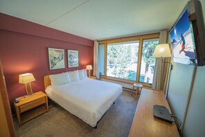 Habitación de hotel con una cama grande, un escritorio con lámpara, televisor y ventana con vista a árboles.