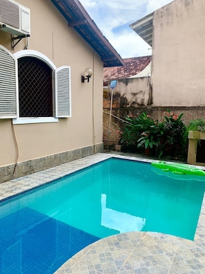 Une piscine dont le bord est recouvert de carrelage, une maison à l’extérieur beige et un toit en tuiles.