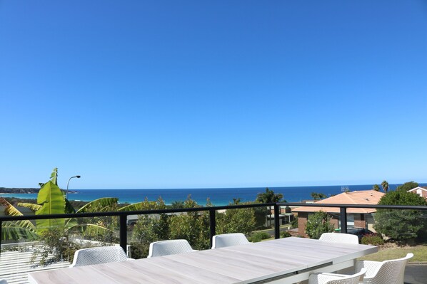 Una terraza con mesa de madera y sillas blancas, con vista al litoral, al océano y a las casas.