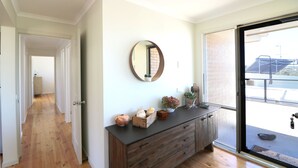 Un couloir doté d’un plancher en bois, une console en bois foncé agrémentée d’objets décoratifs, un miroir rond et une porte-fenêtre coulissante donnant sur un espace extérieur.