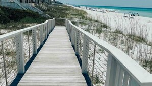 Une promenade en bois menant à une plage, avec des garde-corps blancs, une plage de sable et un ciel bleu clair.