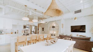 Une cuisine spacieuse avec des meubles blancs, un coin repas comprenant une table et des chaises blanches, et un salon avec un canapé marron et un téléviseur fixé au mur.