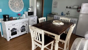 A dining area with a wooden table and white chairs, a white cabinet with drawers, a decorative wall piece, and a kitchen area with a refrigerator and microwave. - Hummingbird Cottage: Cozy Rustic Lakefront Escape at The Pier, Perfect for 7! (Union City)