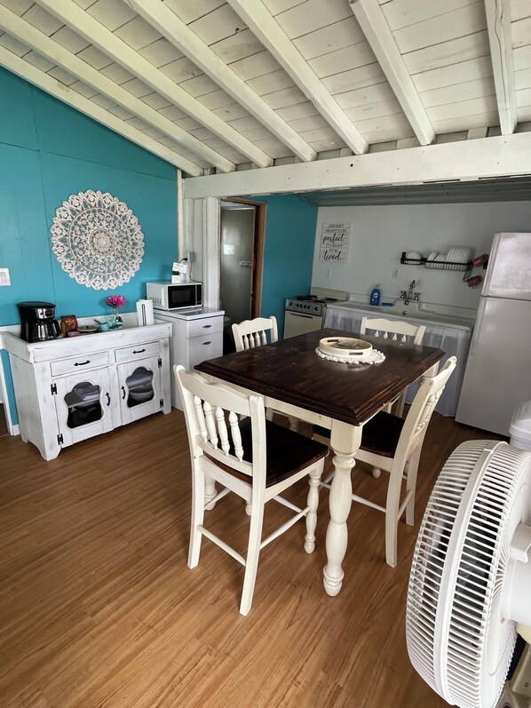 A dining area with a wooden table and white chairs, a white cabinet with drawers, a decorative wall piece, and a kitchen area with a refrigerator and microwave.
