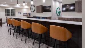 A modern kitchen with a bar area, featuring a large island with stools, pendant lights, a clock, and a TV mounted above the counter. - GrandStay Hotel & Suites Algona IA (Algona)