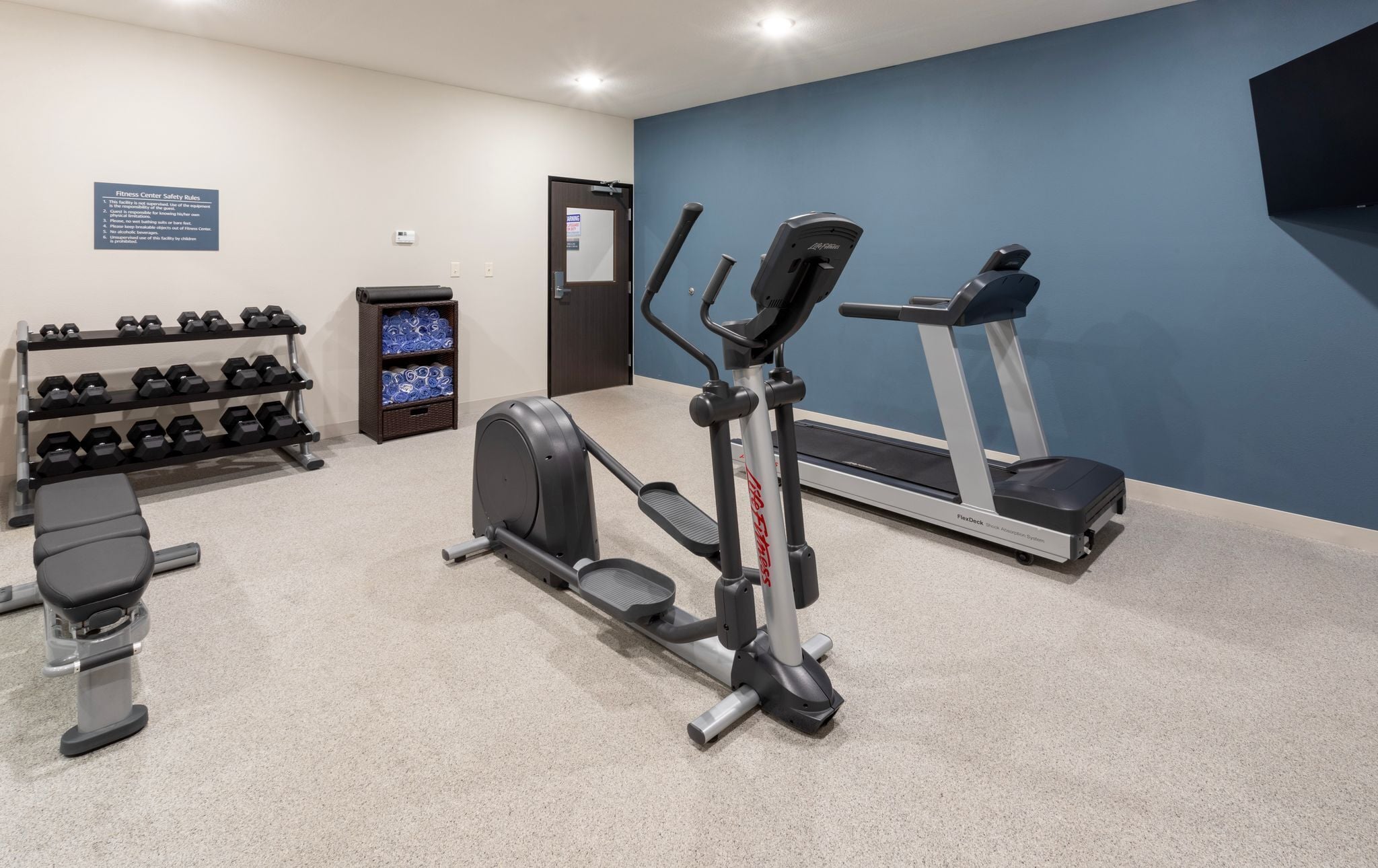 Un gimnasio con cinta de correr, elíptica, mancuernas y banco.