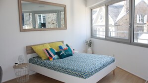 A bedroom with a bed, a round side table, a mirror, and a window with a view of buildings. - Studio de 33 m² en Centre-ville (Saint-Brieuc)