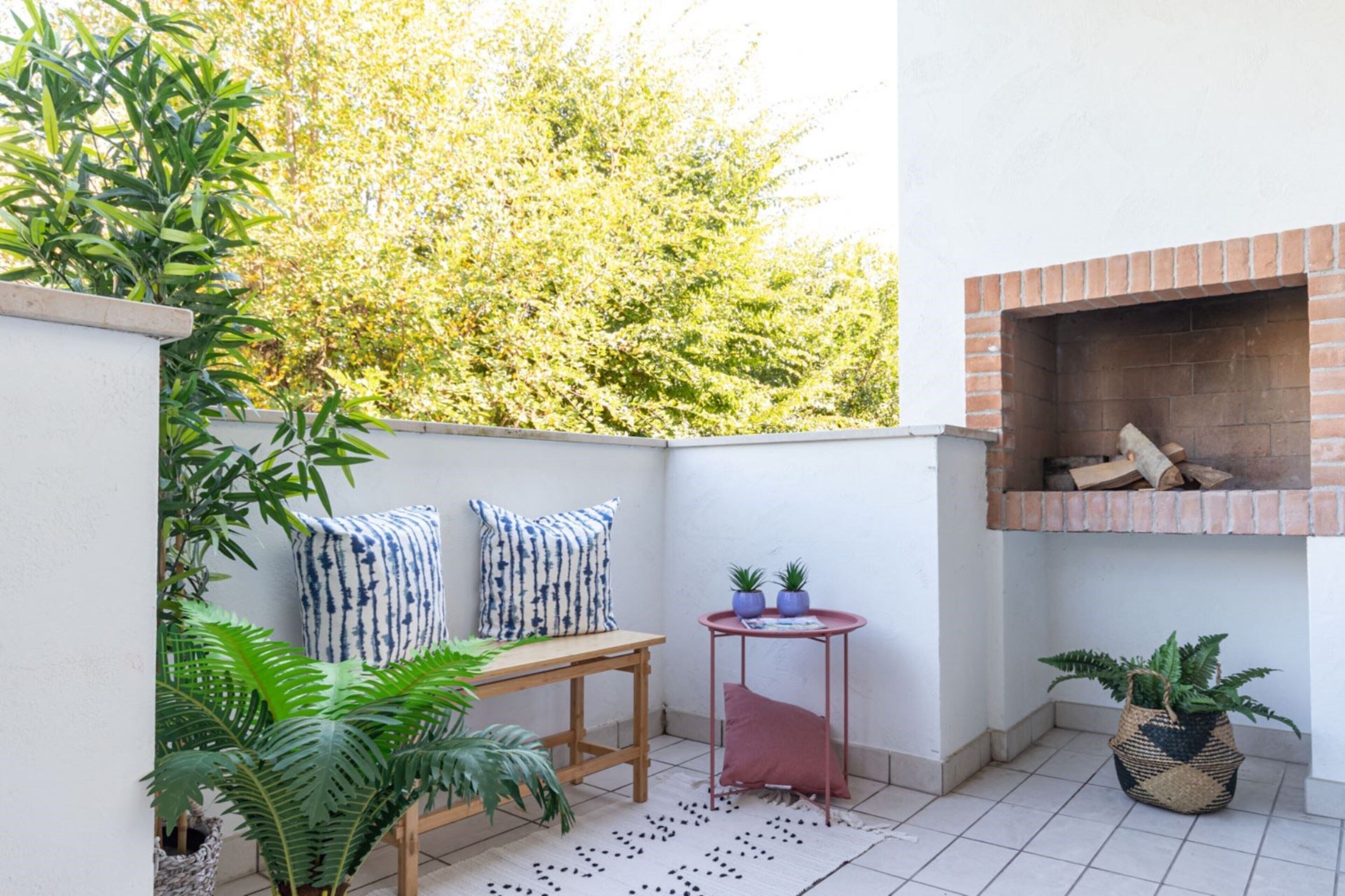 A small patio area with a brick fireplace, a wooden table, two potted plants, and a patterned rug.