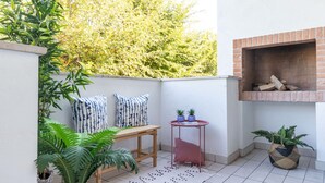 A small patio area with a brick fireplace, a wooden table, two potted plants, and a patterned rug. - Stunning Apartment in a Holiday Village With Pool (San Michele al Tagliamento)