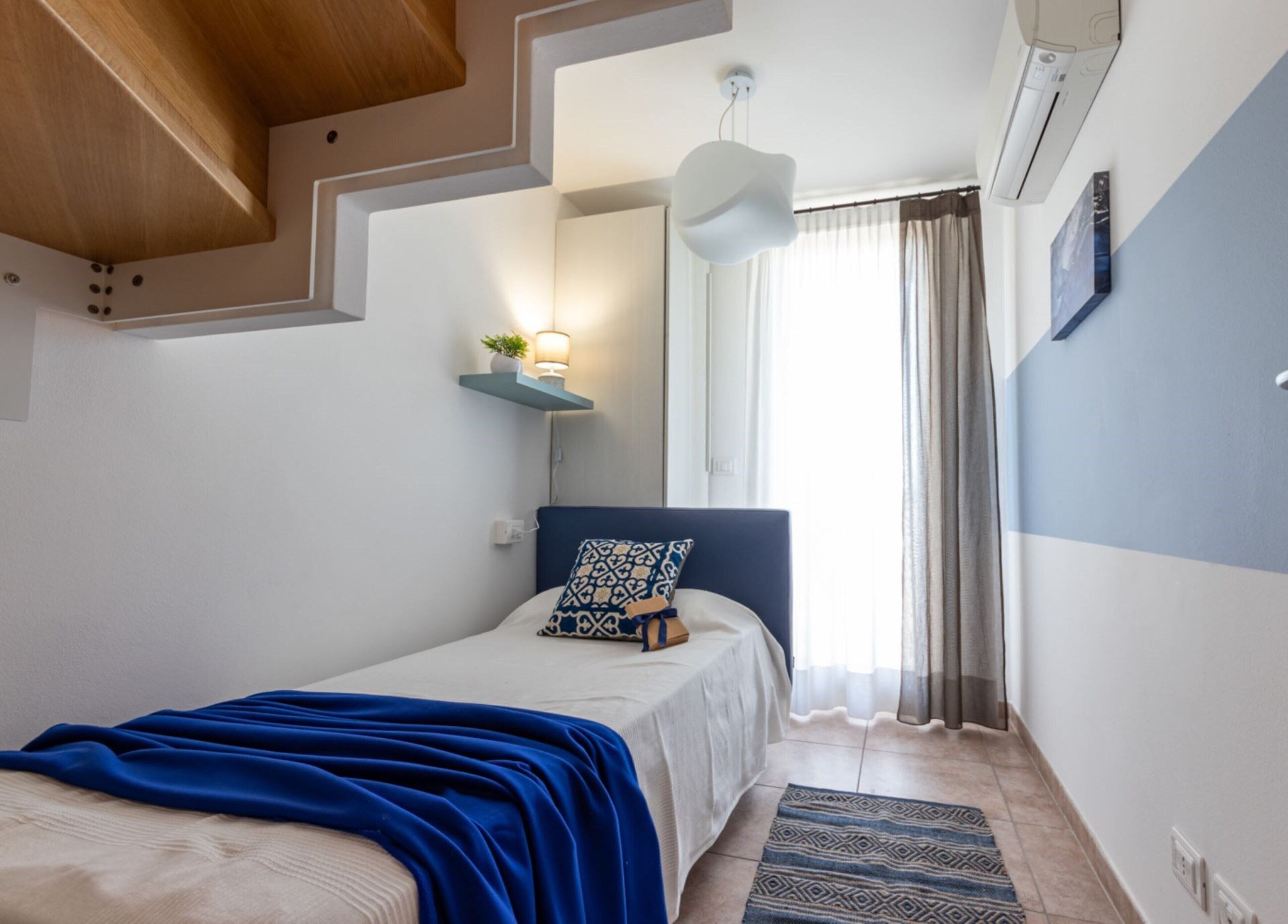 A bedroom with a bed, a bedside table, a wall-mounted shelf with a plant, a hanging light fixture, and a window with curtains.