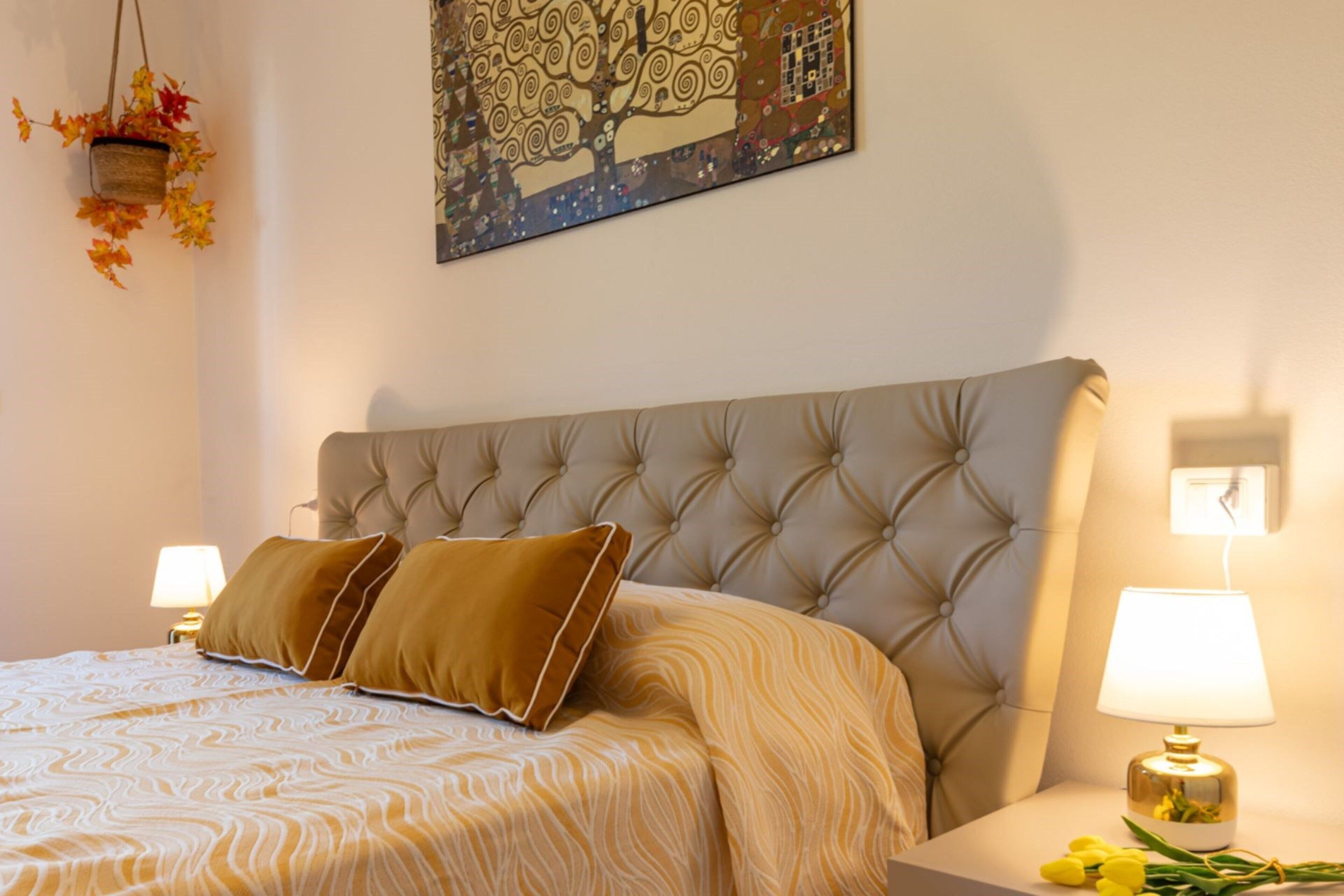 Un dormitorio con cabecera capitonada, dos lámparas de noche, cama con colcha estampada y una planta colgante con hojas otoñales.