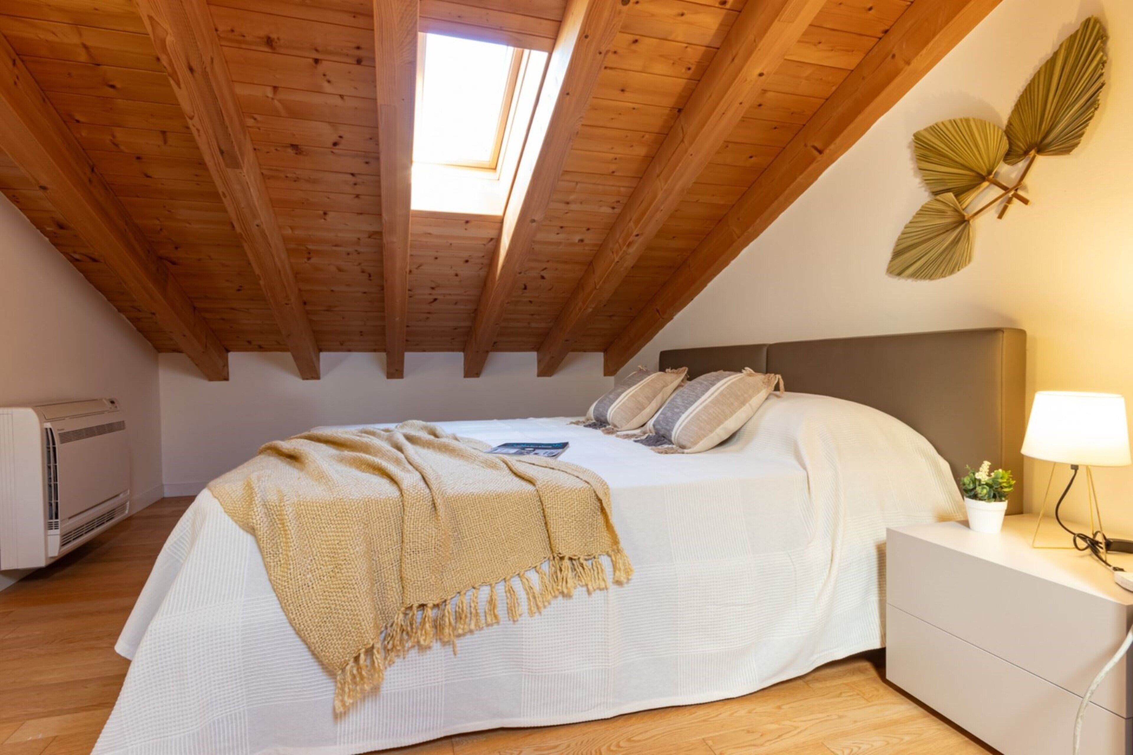Ein Schlafzimmer mit schrägverlegtem Holzboden, einem Bett mit beiger Decke, einer Nachttischlampe und einer Wandpflanze.