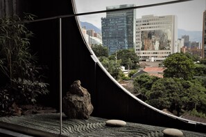 Vista desde una habitación con un separador de vidrio curvo, que presenta un jardín de rocas, una planta de bambú y una pincelada de paisaje urbano con edificios y árboles.