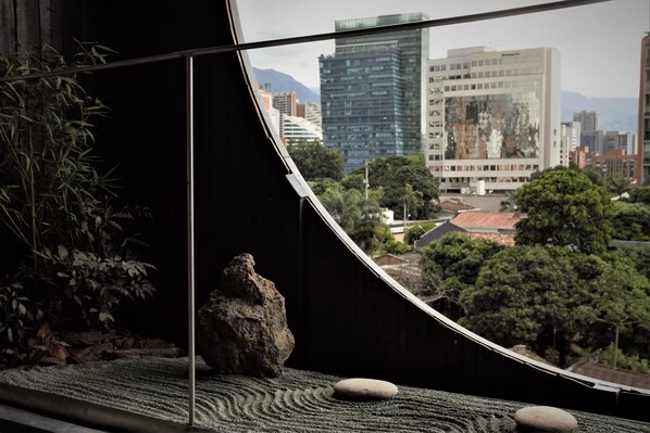 Vue depuis une pièce dotée d’une cloison en verre incurvé, avec un jardin de rochers, un plant de bambou et un aperçu d’un paysage urbain avec des bâtiments et des arbres.