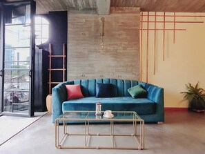 Un salon moderne avec un canapé de couleur turquoise, une table basse en verre et un mur en béton orné d’un motif géométrique.