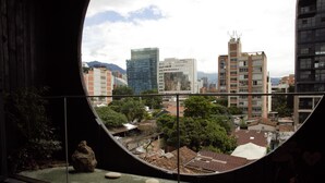 Une vue à travers une fenêtre circulaire sur un paysage urbain, avec des bâtiments modernes, un jardin sur le toit et un grand rocher qui en constitue l’élément central.